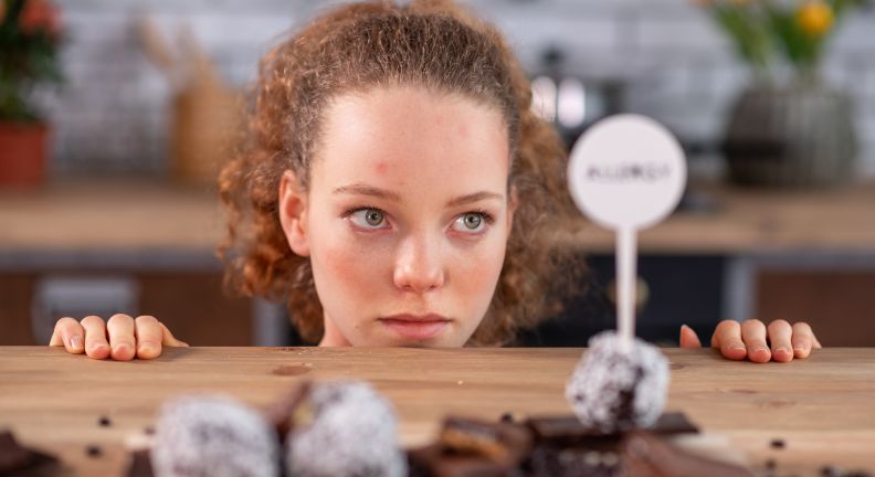 Junge Person schaut über eine Tischkante auf Pralinen mit Kokosraspeln, daneben ein Schild mit der Aufschrift „KUCHEN“