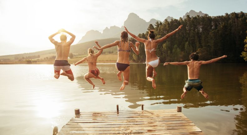 Fünf Personen springen von einem Holzsteg in einen See, im Hintergrund Wälder und Berge.