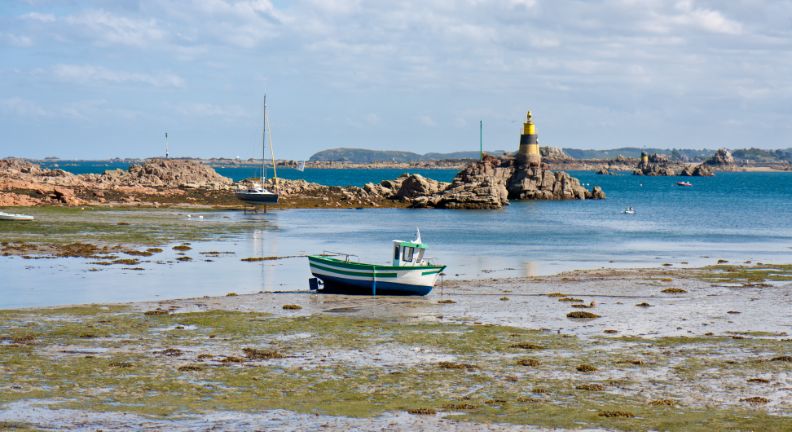 Kleines Boot liegt bei Ebbe auf dem sandigen Meeresgrund, im Hintergrund Felsen, Segelboote und ein Leuchtturm.