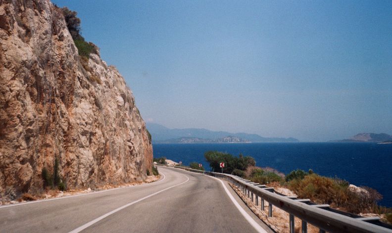 üstenstraße mit Kurve entlang eines Felsens, rechts das Meer und im Hintergrund eine hügelige Landschaft unter wolkenlosem Himmel.