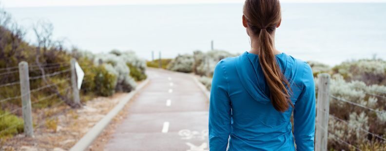 Person steht auf einem Weg am Meer und blickt in Richtung Wasser. Der Weg ist von niedrigen Pflanzen und einem Zaun gesäumt.