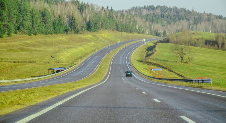 Breite Landstraße mit mehreren Fahrspuren durch eine grüne Hügellandschaft, wenige Autos in der Ferne.