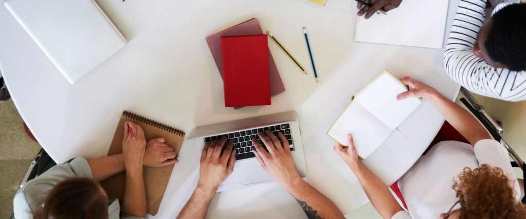 Mehrere Personen sitzen um einen Tisch, mit Laptop, Notizbüchern und Stiften, Ansicht von oben.