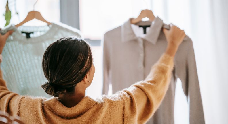 Person hält zwei Kleidungsstücke auf Kleiderbügeln hoch, links ein hellgrüner Pullover, rechts ein graues Hemd.