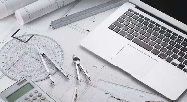 Laptop liegt auf technischen Zeichnungen mit Lineal, Taschenrechner, Zirkel und Geodreieck.