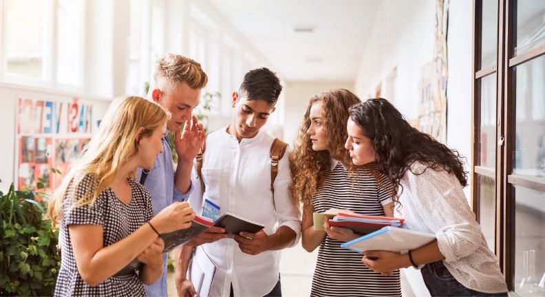 Fünf Jugendliche stehen im Flur und schauen gemeinsam in ein Notizbuch, einige halten Hefte und Bücher.