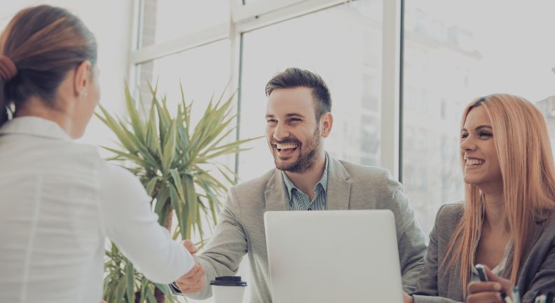 Mann und Frau im Business-Outfit sitzen am Tisch, der Mann schüttelt einer weiteren Person die Hand, alle lächeln.