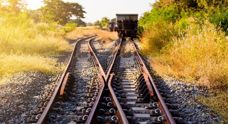 Eine Eisenbahnschiene mit Weichen, auf der ein Wagon abgestellt ist. Im Hintergrund ist eine Naturlandschaft, die idyllisch im Sonnenlicht strahlt.