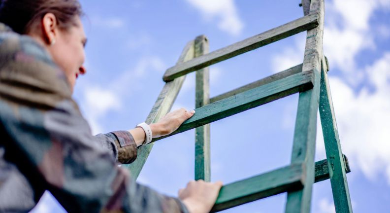 Person greift mit beiden Händen an eine grüne Leiter; im Hintergrund blauer Himmel mit Wolken.