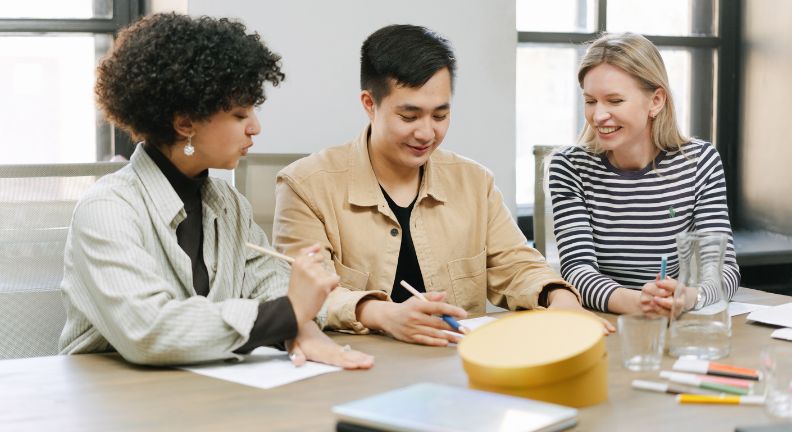 Drei Personen sitzen am Tisch und schreiben auf Papier; auf dem Tisch liegen Stifte, Gläser, Karaffe und eine runde Schachtel.