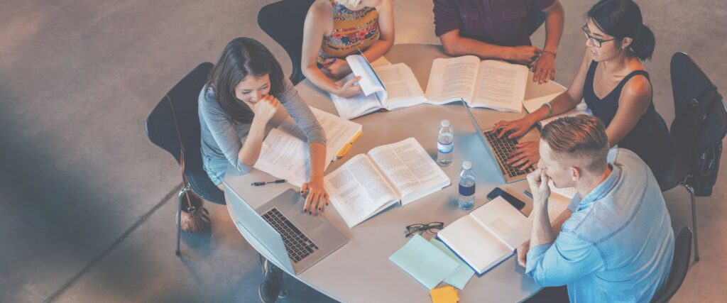 Vier Personen an rundem Tisch mit Laptops, Büchern und Notizen; zwei Wasserflaschen und Smartphone auf dem Tisch.
