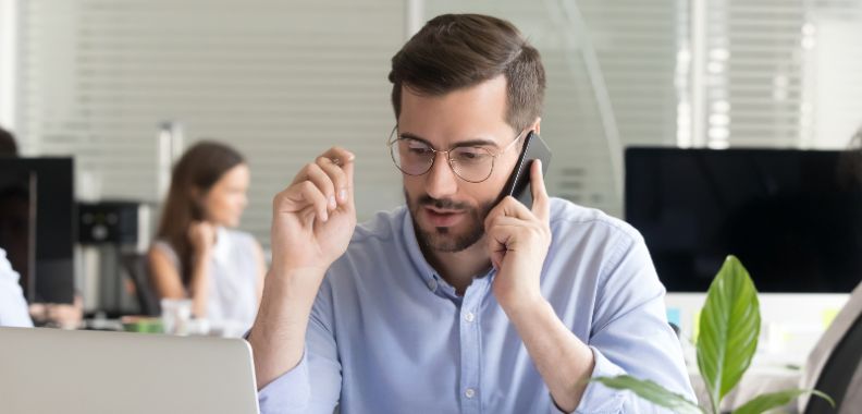 Mann sitzt am Schreibtisch, telefoniert und blickt auf Laptop; im Hintergrund Büro mit weiteren Personen und Bildschirmen.