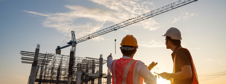 Zwei Personen mit Schutzhelmen und Warnwesten zeigen auf eine Baustelle mit Baukran und Stahlgerüst bei tief stehender Sonne.