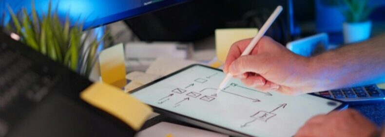 Hand zeichnet mit Eingabestift auf Tablet; im Hintergrund Monitore mit Programmcode und Schreibtisch mit Haftnotizen.