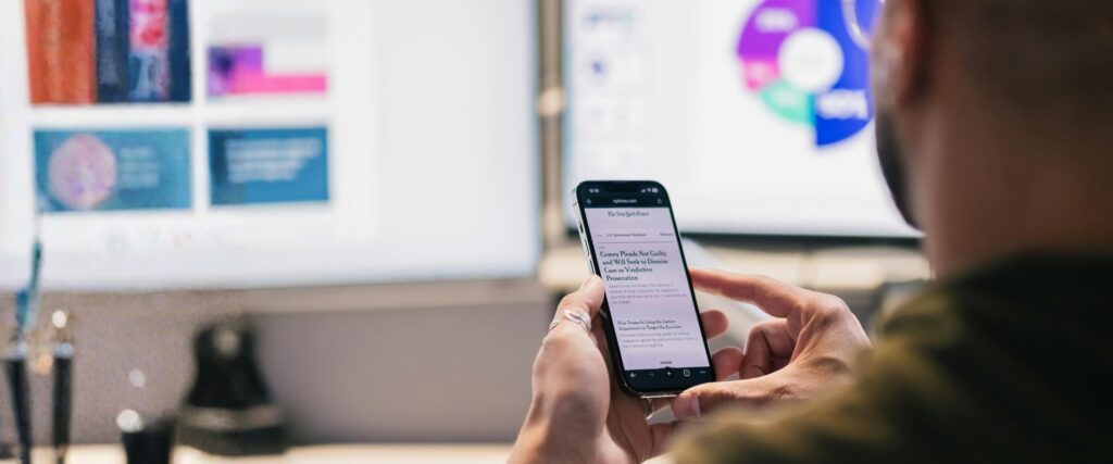 Person hält ein Smartphone mit geöffneter Nachrichtenseite; im Hintergrund unscharfe Monitore mit Grafiken und Diagrammen.