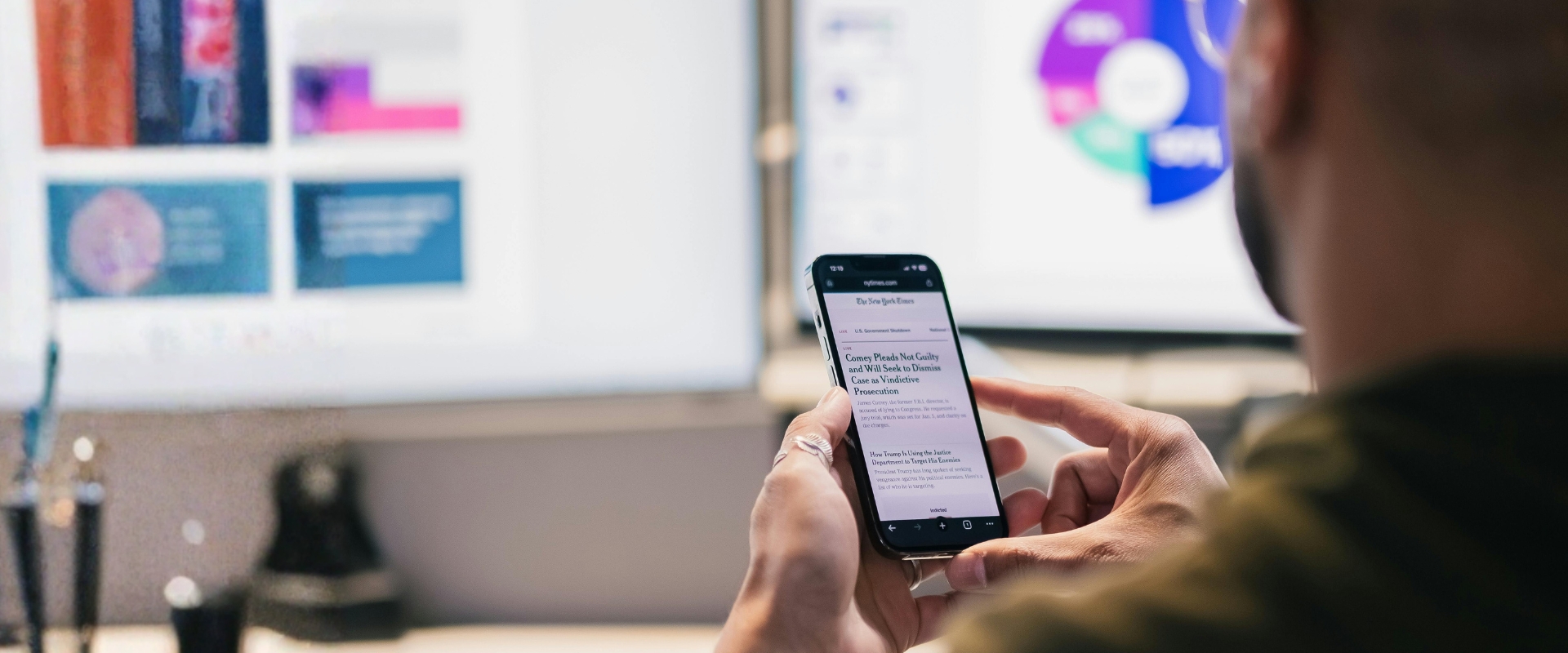 Person hält ein Smartphone mit geöffneter Nachrichtenseite; im Hintergrund unscharfe Monitore mit Grafiken und Diagrammen.