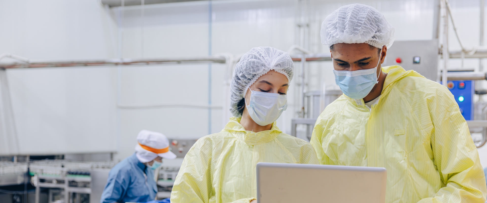 Drei Personen mit Haarnetzen, Mund-Nasen-Masken und Schutzkleidung stehen in einer Produktionshalle; zwei blicken auf einen Laptop.